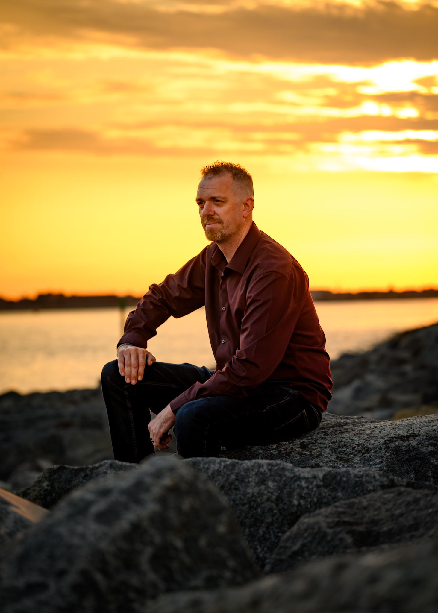 Seated environmental portrait among rocks beneath a dramatic golden sky.