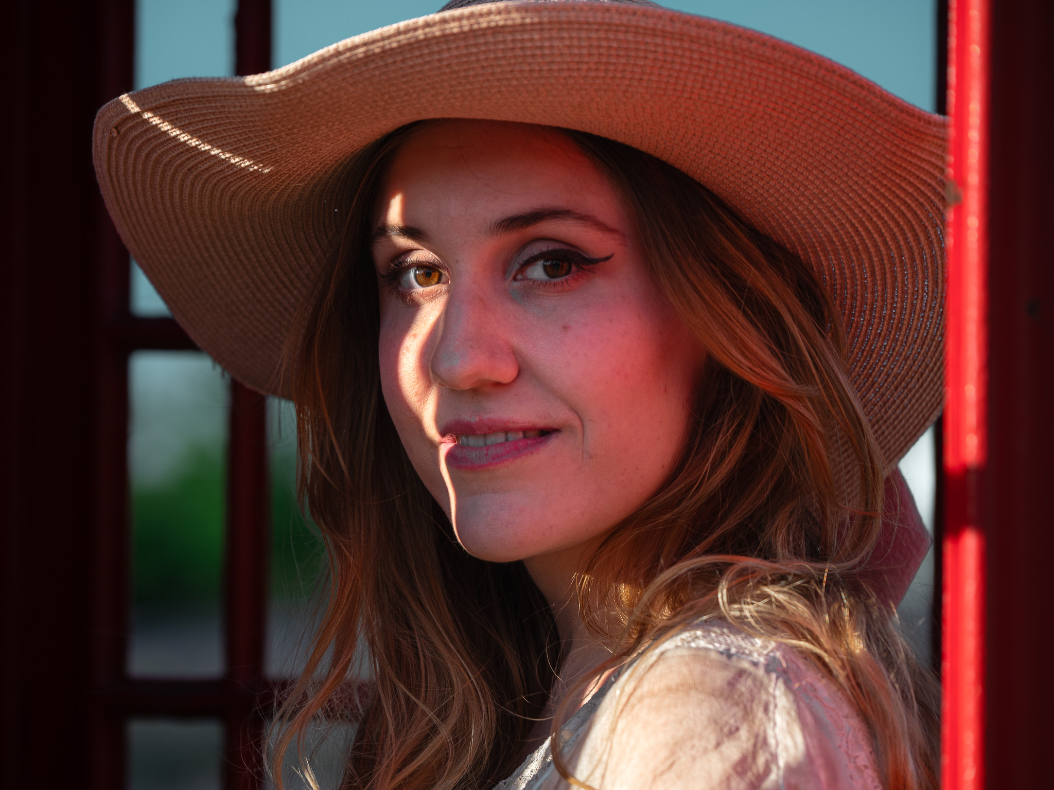 Warm close portrait of Andreea in a wide-brimmed hat, framed by red-painted wood and a stripe of sunlight